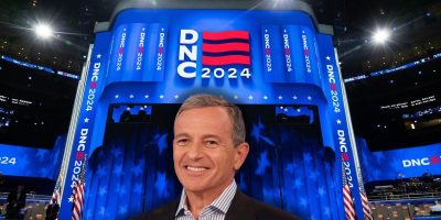 Disney CEO Bob Iger, wearing a dark suit and light shirt, stands smiling in front of a large, brightly lit stage with the sign "DNC 2024" displayed prominently in blue and red. The stage, reminiscent of Disney magic, is set up with multiple "DNC 2024" banners and American flags.