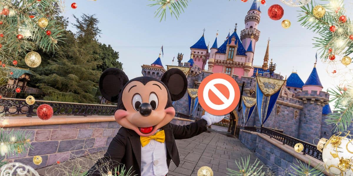 A person in a Mickey Mouse costume stands in front of a castle at Disneyland, holding a "No Entry" sign. The scene is festively decorated with Christmas ornaments and greenery. The sky is clear and blue, and the castle’s blue rooftops stand out against the background.