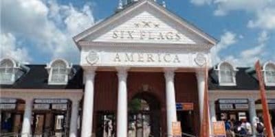 Entrance of Six Flags America featuring a large building with white pillars and a triangular pediment displaying the park's name. Ticket booths are visible below, with employees assisting eager visitors, surrounded by signs and people. The sky is partly cloudy.