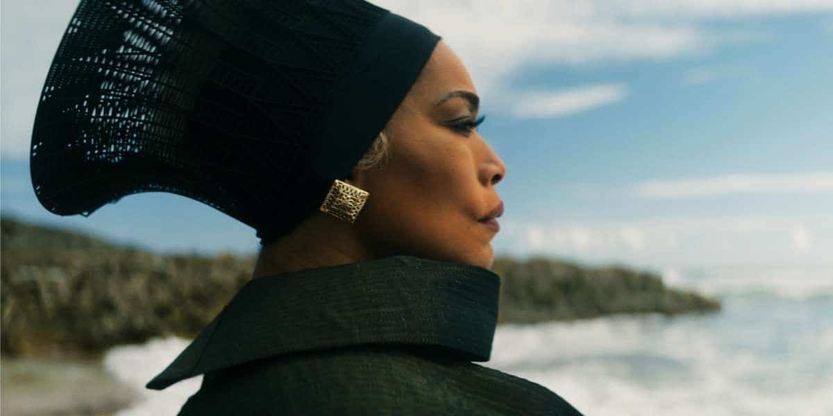 A person stands by the sea in profile view, facing the horizon. They wear a structured, black headdress and matching outfit with a high collar, paired with gold, square earrings. The sea and sky are visible in the background, showing a calm and serene setting.