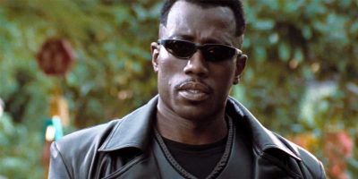 Blade (Wesley Snipes) wearing dark sunglasses and a black leather jacket stands outdoors, looking serious. The background is blurred with greenery, creating a contrast with the person's dark attire, reminiscent of Wesley Snipes in the Blade reboot within the MCU.