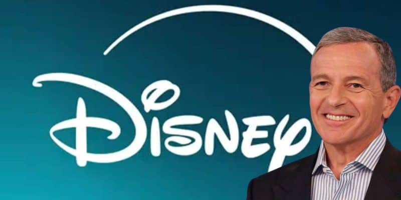 A man in a suit is smiling in the foreground. Behind him, there is a large white Disney logo with a blue background.