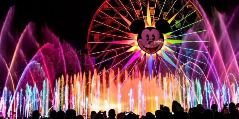 Brightly colored water fountains light up at night during World of Color beneath a Ferris wheel with a large Mickey Mouse face in the center at Disney California Adventure Park.