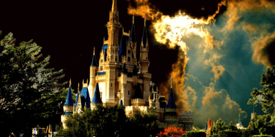 A majestic castle with blue rooftops and golden spires sits against a dramatic sky where dark clouds and bright sunlight create a striking contrast. Lush green foliage surrounds the castle, reminiscent of the Magic Kingdom, adding to the fairytale-like atmosphere.