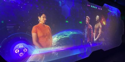 A futuristic display features a woman in an orange outfit on the left and two men in cosmic attire on the right. They appear to be in a space-themed virtual environment, with cosmic visuals and the Earth in the background. A hexagonal interface overlays the image, reminiscent of a Disney Coaster adventure.