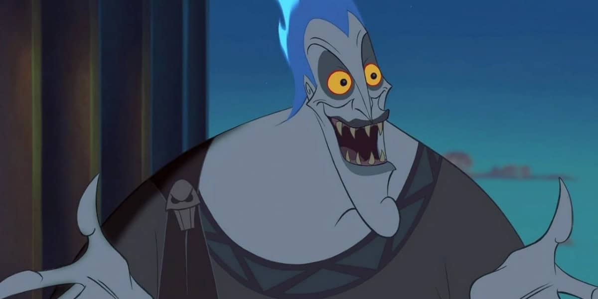 An animated character with gray skin, blue flaming hair, and yellow eyes, wearing a dark toga-like outfit, grins widely with arms outstretched. The character has exaggerated features and a mischievous expression. The background shows a dimly lit environment.