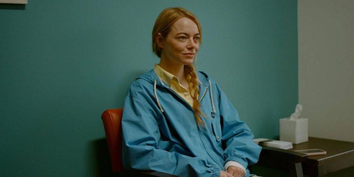 A woman with light hair is sitting on a red chair against a teal wall, wearing a blue jacket over a yellow shirt. She looks ahead with a slight smile. There's a Coming Soon poster for Disney+ on the table to her left.