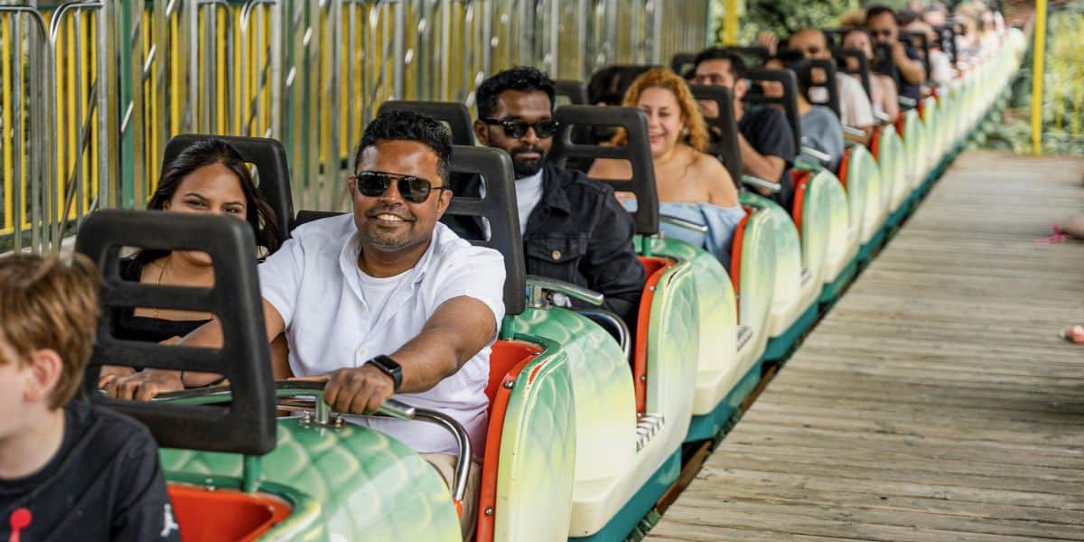 People are seated in two-person rows on a roller coaster ride, smiling and wearing casual clothing. The ride appears to be in a theme park setting with green and yellow barriers on the side. The atmosphere is joyful and lively.