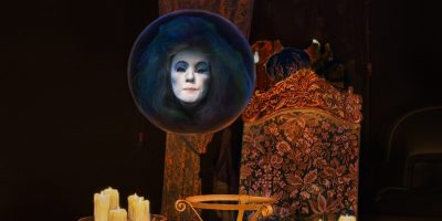 A spectral face with dramatic makeup appears in a glowing crystal ball in a dimly lit room. The scene includes a richly upholstered chair, several lit candles, and dark drapery, evoking a mystical and eerie atmosphere.