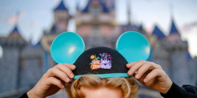 Close-up of a person wearing a black hat with blue mouse ears and a colorful "Disneyland Resort" logo, holding the brim of the hat with both hands. The background shows a blurred view of a castle with blue rooftops, capturing the magic of D23: The Ultimate Disney Fan Event.