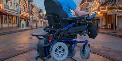 A person wearing a blue shirt is seen from behind using a motorized wheelchair on an empty street lined with buildings adorned with lights, reminiscent of the enchanting ambiance created for Disney guests. The atmosphere suggests a festive or amusement park setting.