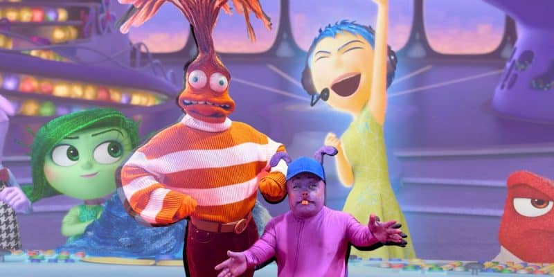 A vivid scene featuring characters from the animated movie "Inside Out." Joy is cheerfully throwing her arms up, while Disgust, Anger, and Fear appear in the background. A person in a creative Disney cosplay stands in the foreground, resembling a hybrid fruit and vegetable figure.