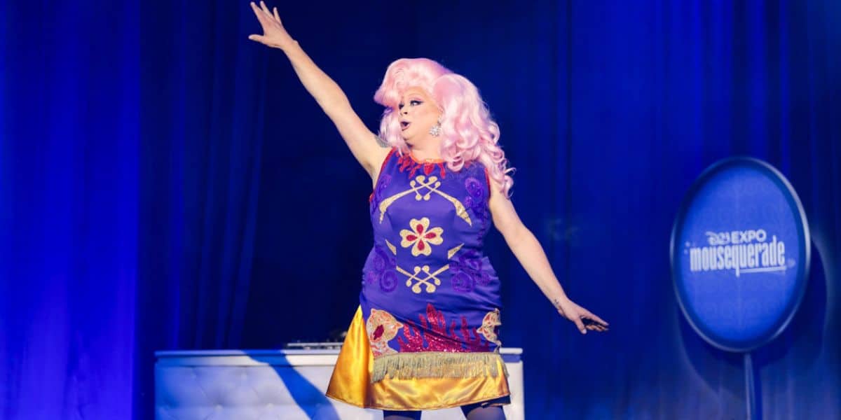 Nina West with bright pink hair is on stage, raising one arm and wearing a colorful purple dress with floral patterns. The background features blue curtains and a sign that reads "D23 Expo Mousequerade." Channeling the magic of Disney Drag Queens, the stage also features a white DJ booth.