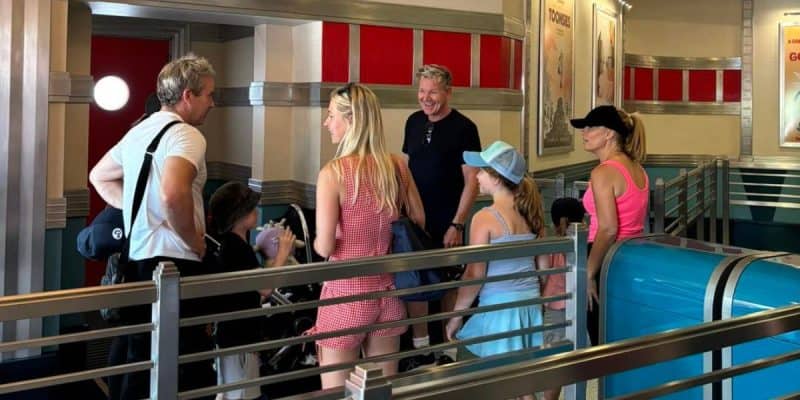 A group of people, including men, women, and children, and Gordon Ramsay, stand together inside a retro-styled building with metallic and red decor. They appear to be in conversation and look casual and happy, much like celebrities at Disney. The group is standing near railings and a blue trash bin.