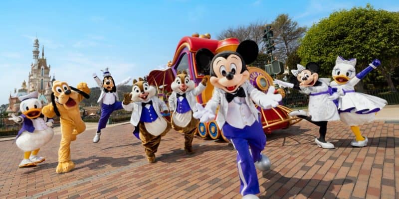 Disney characters, including Mickey Mouse, Minnie Mouse, Donald Duck, Daisy Duck, Goofy, and Pluto, are dressed in festive attire and marching energetically in front of a red parade float. Cinderella’s Castle is visible in the background under a clear blue sky inside Hong Kong Disneyland.