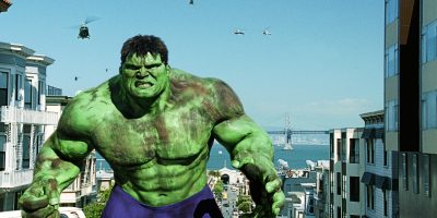 The Incredible Hulk, a large green muscular figure, stands in the middle of a city street with buildings on both sides. Helicopters are flying in the sky, and a bridge over water is visible in the background.