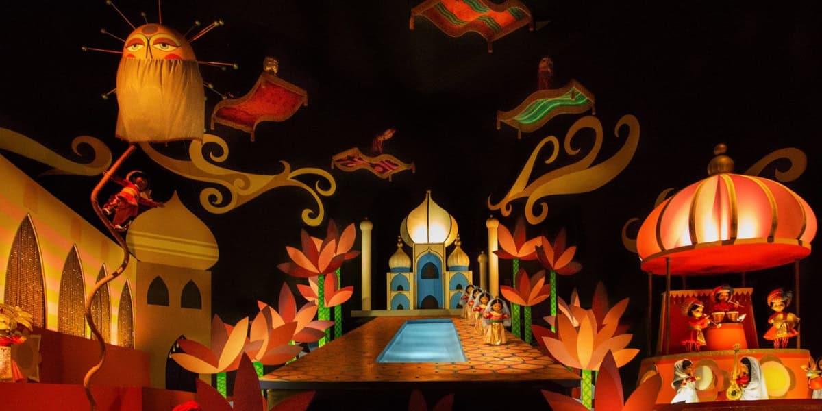 A vibrant indoor scene depicting an Arabian Nights theme with animated figures. There are ornate palaces, magic carpets floating in the air, and various brightly colored decorations. A pool leads to a central golden pavilion surrounded by stylized trees.