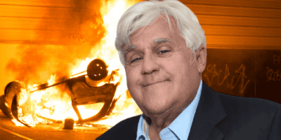 A gray-haired man, reminiscent of Jay Leno, is smiling in the foreground, dressed in a suit. In the background, a car is overturned and engulfed in flames, with a graffiti-covered wall behind it.
