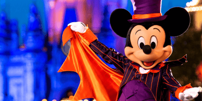 Mickey Mouse dressed as a vampire in a black and purple striped suit with an orange cape, celebrating Halloween. The background features a brightly lit castle at night with blurred lights adding to the festive atmosphere.