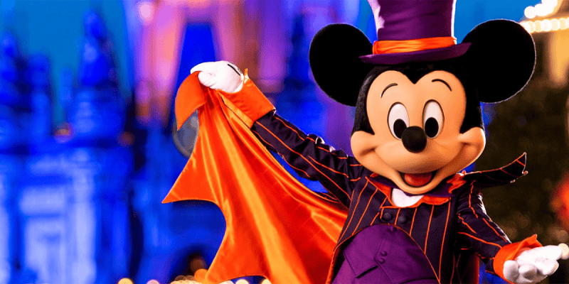 Mickey Mouse dressed as a vampire in a black and purple striped suit with an orange cape, celebrating Halloween. The background features a brightly lit castle at night with blurred lights adding to the festive atmosphere.