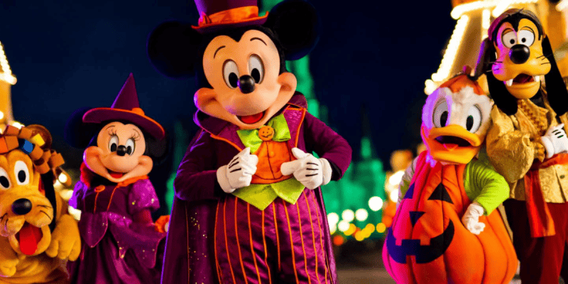 A group of Disney characters dressed in Halloween costumes, including Mickey Mouse in a top hat and cape, Minnie Mouse in a witch outfit, Donald Duck in a pumpkin costume, Goofy in a skeleton costume, and Pluto with a festive collar, standing together at night.