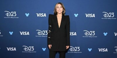 Miley Cyrus poses in a black suit against a blue D23 backdrop.