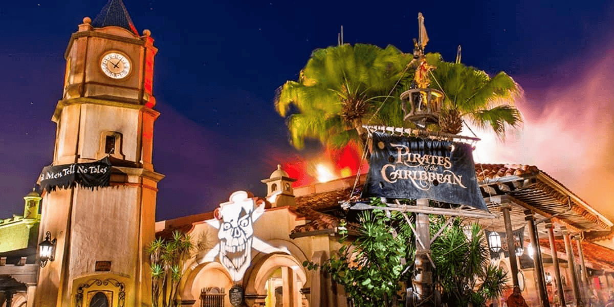 A brightly lit entrance to the "Pirates of the Caribbean" theme park attraction at night, featuring a clock tower, a pirate skull and crossbones sign with the ride's name, and lush palm trees. The colorful sky adds to the vibrant and adventurous atmosphere.