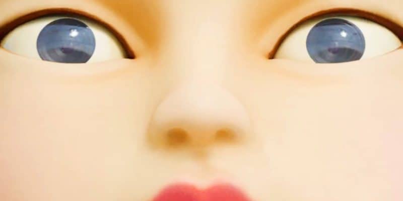 Close-up image of a doll's face with wide-open blue eyes, a small nose, and bright red lips. The doll's skin is smooth and pale, and the expression appears surprised or shocked.