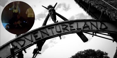 A large metal archway with the word "Adventureland" displayed prominently in a decorative, tribal-inspired font. In the top left, a circular inset reveals a dark scene with three individuals—two standing and one on the ground—hinting at an unsettling encounter that nearly shut down the area.