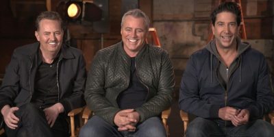 Three men, including Matthew Perry and Matt LeBlanc from Friends, are sitting on director's chairs, smiling at the camera. All three wear casual clothes, such as shirts and jackets. The background is a set with studio lights and equipment, giving a behind-the-scenes feel.