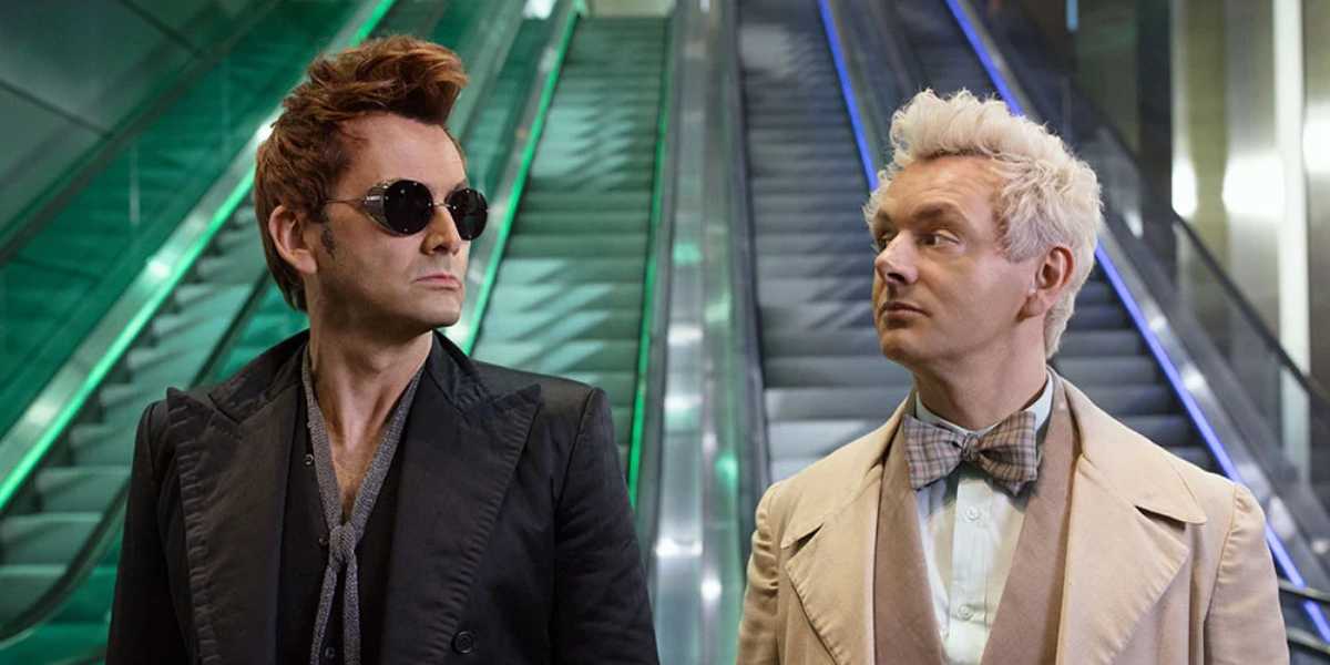 Two men stand in front of an escalator. The man on the left has spiky red hair, dark sunglasses, and wears an all-black outfit. The man on the right has short white hair, and wears a beige coat with a bowtie. Both are looking at each other with neutral expressions, as if in a scene from an Amazon show.