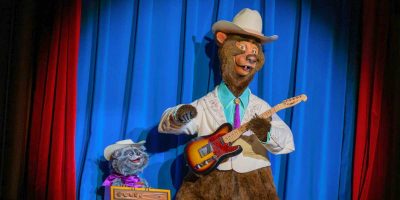 A bear animatronic, reminiscent of an iconic Disney Parks character, dressed in a white cowboy hat and suit plays an electric guitar on a stage with a blue curtain background. Beside it, a smaller bear in a cowgirl hat and purple outfit sits with a box. Both figures appear whimsical and entertaining.