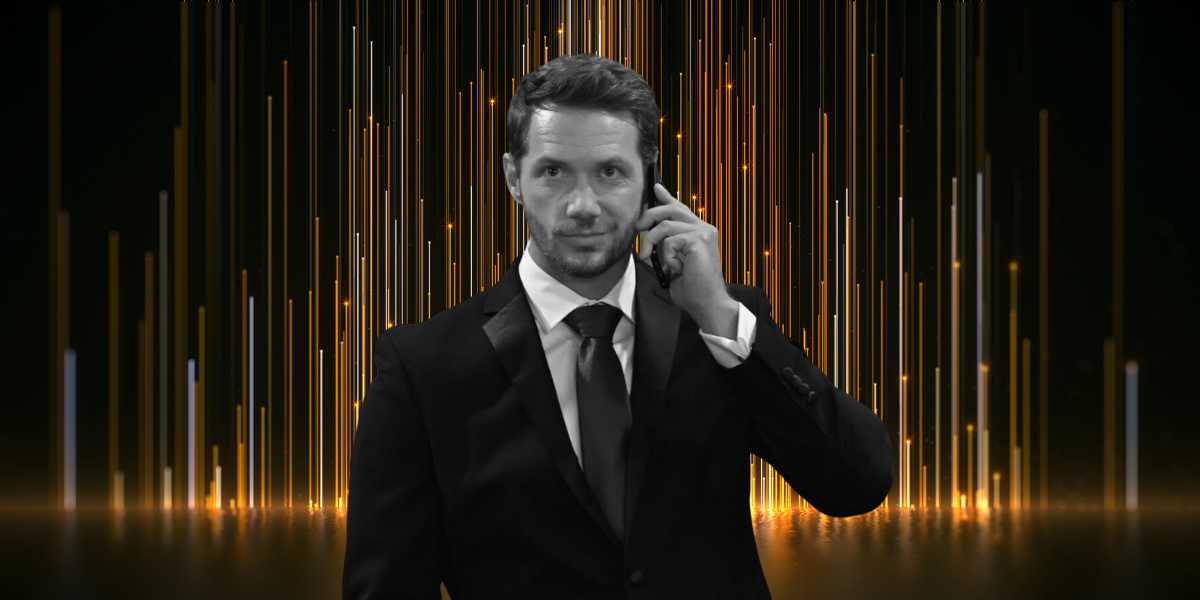 A man in a black suit and tie holds a phone to his ear. He has short hair and a serious expression. The background features vertical, glowing golden lines on a dark backdrop, creating a dynamic and futuristic atmosphere, reminiscent of an awards show tribute that left viewers shocked.
