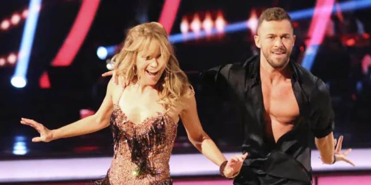 Two dance contestants energetically perform on stage under colorful, dynamic lighting. The female dancer, perhaps seeking a joyful escape after a brutal assault, is wearing a sparkling, fringed dress. The male dancer is in a black outfit with an open shirt. Both are smiling and engaged in a lively dance routine.