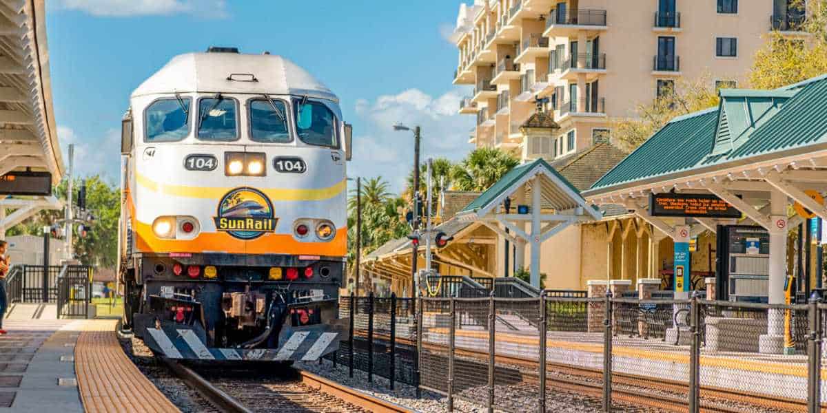 A SunRail train, numbered 104, approaches a station platform. The station features covered seating areas, informational displays, and nearby buildings, including a multi-story residential structure. The clear sky and abundant sunlight provide excellent visibility as passengers await their ride with anticipation.