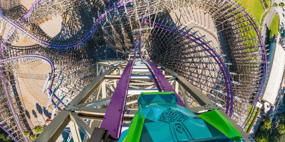 Aerial view from the top of a steep, colorful roller coaster track with twists and turns at a Florida theme park, surrounded by intricate wooden support structures. The vibrant green and purple tracks descend steeply, highlighting the thrilling drop ahead. Green spaces and pathways are visible below as park operations buzz with activity.