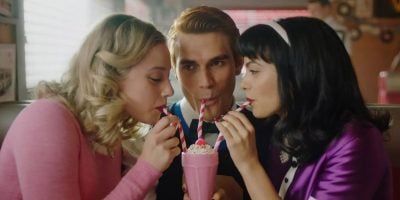Three people are sitting in a diner booth and sharing a milkshake. The person on the left has blonde hair and is wearing a pink sweater, the person in the middle has red hair and is wearing a blue and black shirt, and the person on the right has dark hair and is wearing a purple jacket.