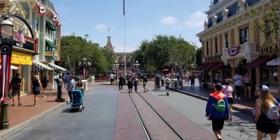 A lively scene at Disneyland's Main Street U.S.A. with visitors walking along the brick-paved road. The street is lined with themed shops adorned with patriotic décor, including banners and American flags. A central flagpole stands tall in the distance.