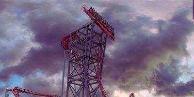 A vibrant, towering roller coaster inside Cedar Point titled Siren's Curse looms against a dramatic sky filled with dark, swirling clouds in North America. The steep drop is filled with riders holding on, facing an intense descent. Known as the fastest coaster, its red tracks and metallic structure contrast with the tumultuous backdrop.