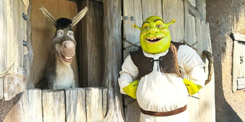 Animated characters Shrek and Donkey from the "Shrek" series are seen at a rustic wooden window. Shrek, a green ogre, stands outside wearing a white tunic and brown vest, smiling widely. Donkey leans out of the window, also smiling. The setting is whimsical and cottage-like.