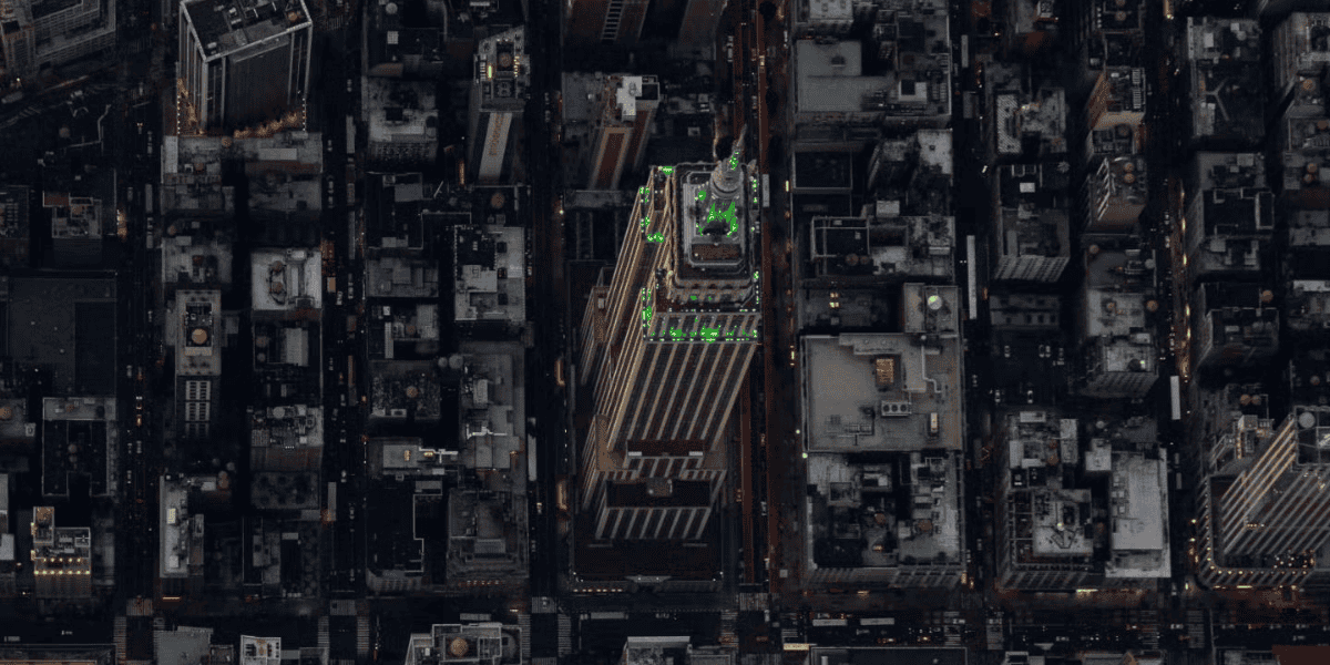Aerial view of a New York cityscape at dusk, featuring a prominent skyscraper with green lit decorations on its upper levels, reminiscent of the Empire State Building, surrounded by other smaller buildings. The streets are filled with vehicular traffic and illuminated by street lights.