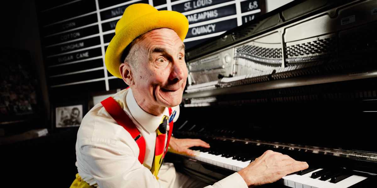 A man with a yellow hat and colorful suspenders, reminiscent of Yehaa Bob, enthusiastically plays the piano. He is smiling and looking upwards, with musical sheet mechanisms visible inside the open piano lid. A black signboard with white text is partially seen in the background.