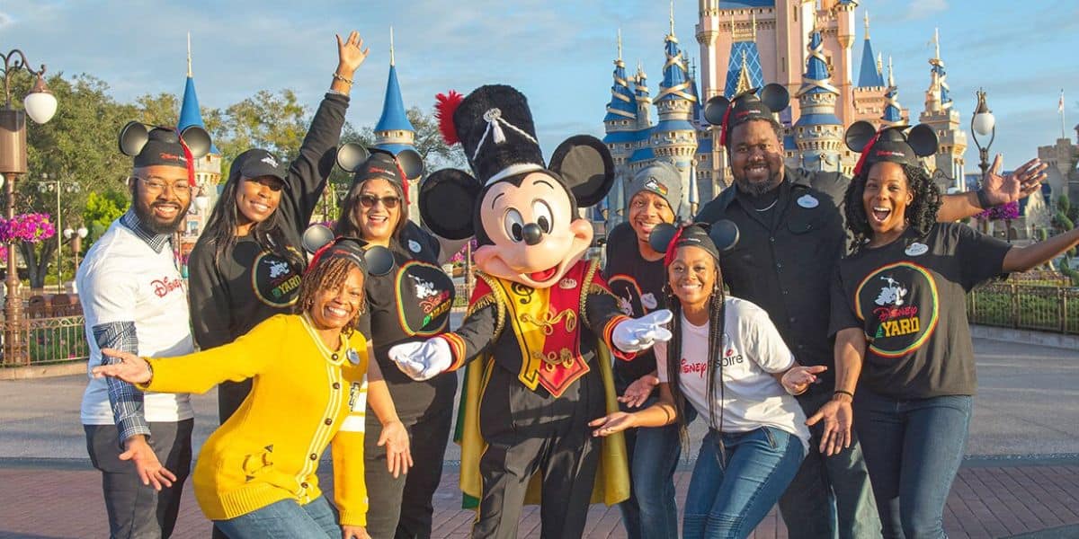 A group of smiling adults wearing Mickey Mouse ears, all part of the Disney College Program, pose with a person dressed as Mickey Mouse in front of a castle at a theme park. The group is making joyful gestures, and the castle in the background is decorated with blue and gold accents.