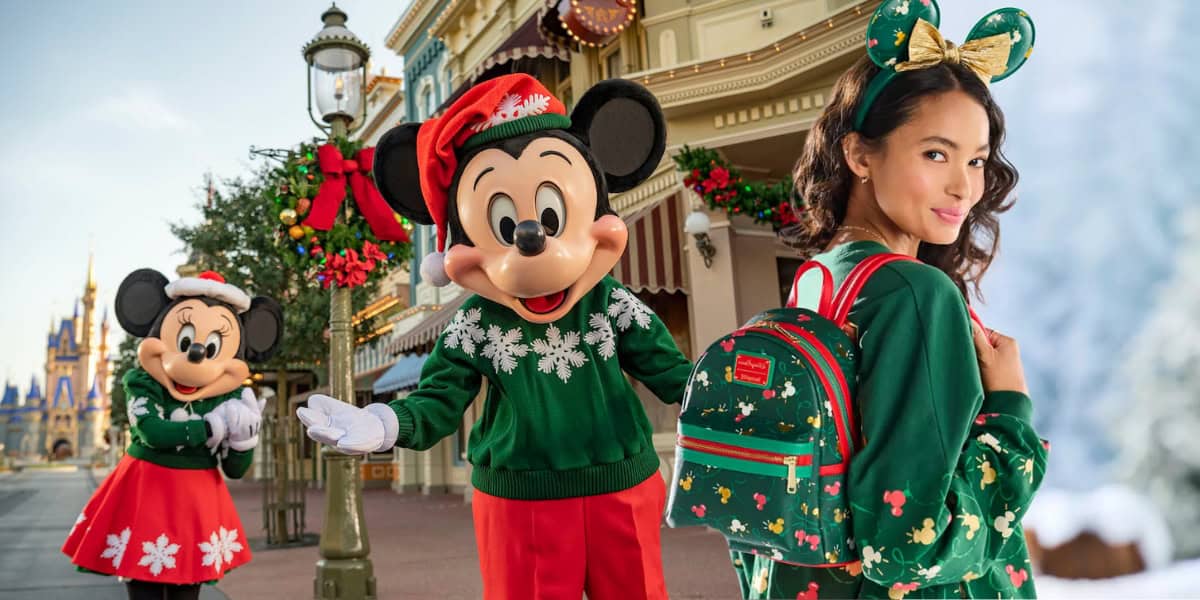 A person with Mickey Mouse ears headband and a green holiday-themed outfit from the Christmas Collection stands on a decorated street with Mickey and Minnie Mouse characters dressed in festive attire near a building adorned with colorful, holiday decorations.