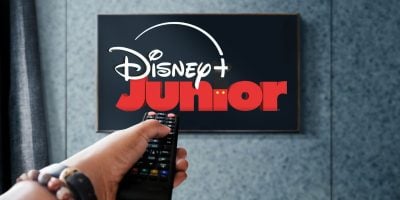 A remote control is pointing it at a television screen displaying the Disney+ Junior logo against a blue-gray wall background. Disney streaming will soon feature a 24/7 Disney Junior channel.