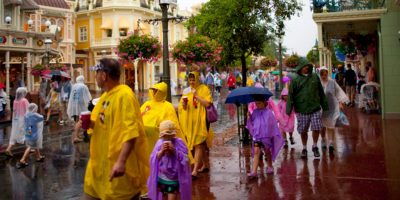A bustling street scene at Disney World on a rainy day. Visitors are wearing colorful rain ponchos and holding umbrellas as they walk past charming, whimsical buildings. Flower baskets hang from lampposts, adding a touch of color to the wet surroundings despite a tornado watch in effect.