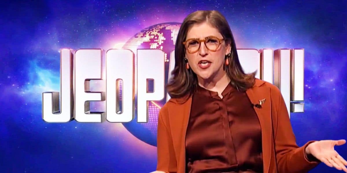 Mayim Bialik speaks animatedly while standing in front of a "Jeopardy!" logo.