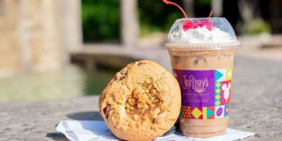 A large chocolate chip cookie sits on a white napkin next to an iced coffee topped with whipped cream and cherries, in a clear plastic cup with colorful branding from Joffrey's Coffee & Tea Company’s Specialty Coffee Collection. The background is slightly blurred with a hint of greenery.