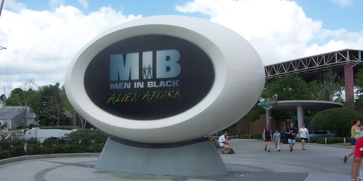 A large elliptical sign with "MIB Men In Black Alien Attack" is displayed outdoors against a blue sky with scattered clouds. People walk near Universal's MIB Attraction sign, and trees and a structure with a roof are visible in the background.