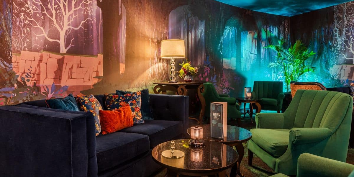 Discover the enchanting new lounge at Disney Hotel, featuring a cozy dark blue sofa with colorful cushions and two green armchairs around a round glass table. A tall lamp and lush plants enhance the warm ambiance against a mystical forest-themed mural.